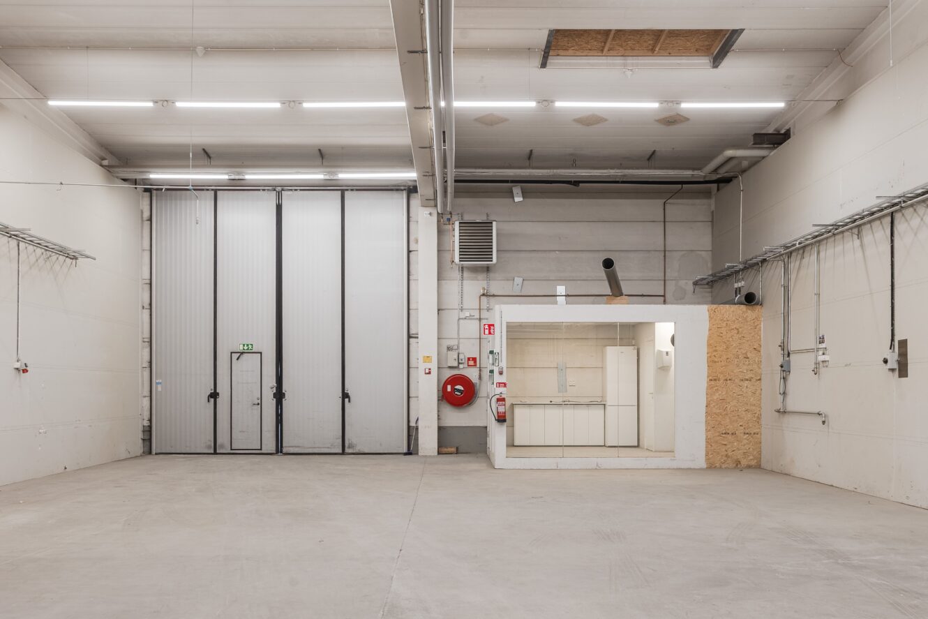 Ett stort tomt industrilager med högt i tak, ett litet slutet kontorsutrymme, betonggolv och en stor garageport i metall.