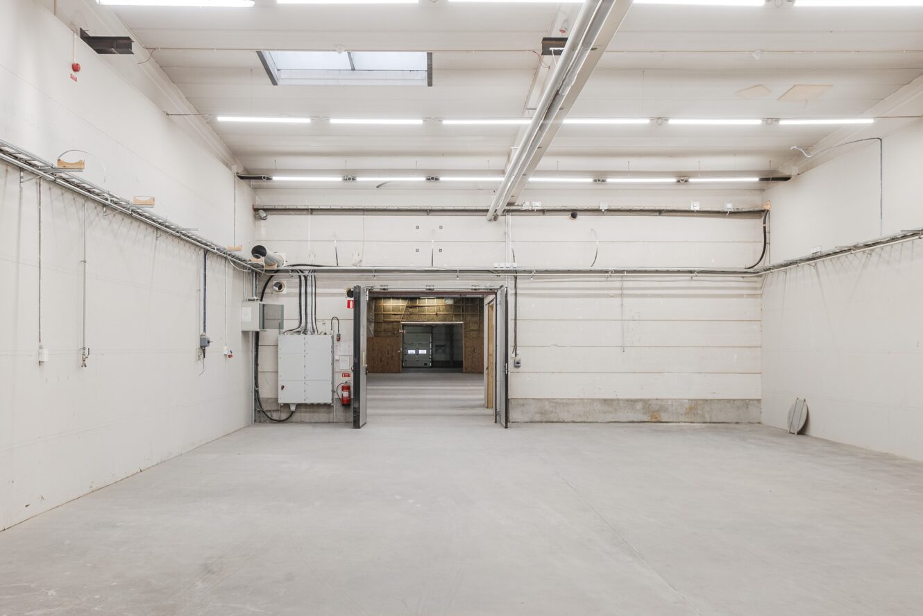 Tomt industrilager med vita väggar, synliga rör och betonggolv; stor öppen dörröppning leder till en annan del i bakgrunden.