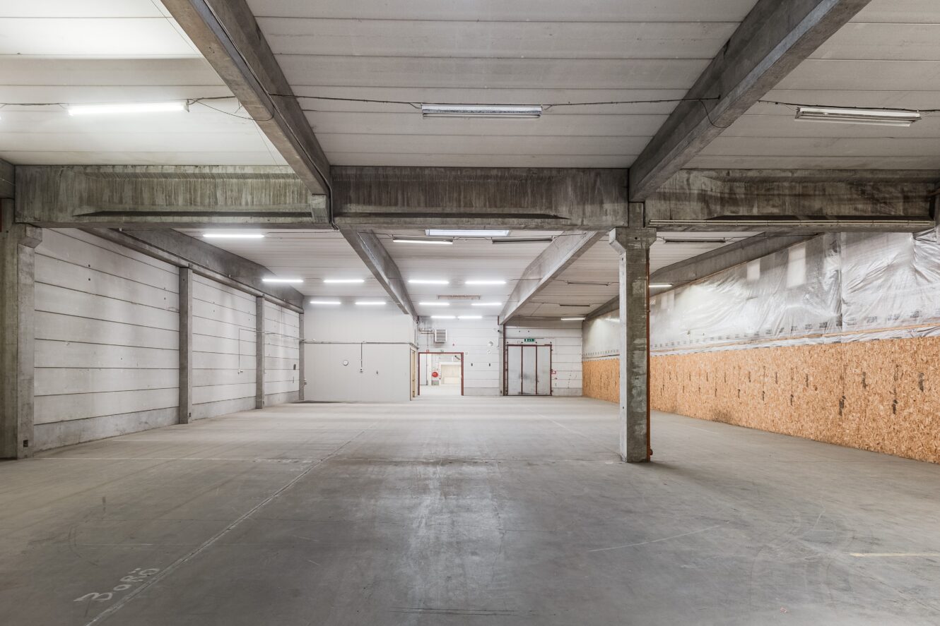 Rymlig, tom industrilokal med betonggolv, högt i tak, synliga bjälkar och lysrörsbelysning. En sidovägg är delvis täckt med träpaneler.