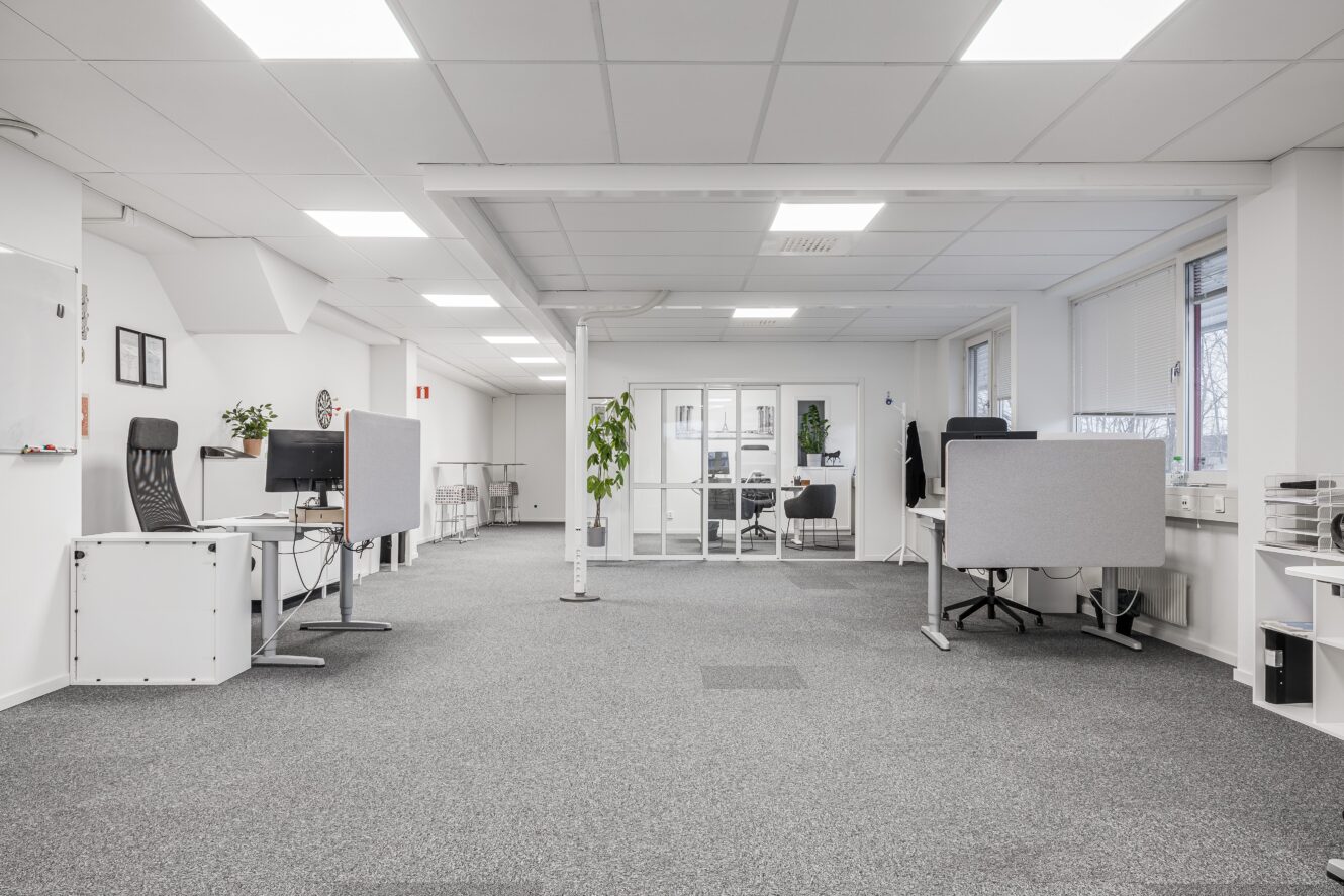 Ett modernt öppet kontorslandskap med grå heltäckningsmatta, flera skrivbord, kontorsstolar, skiljeväggar, stora fönster, vita väggar och inomhusväxter.