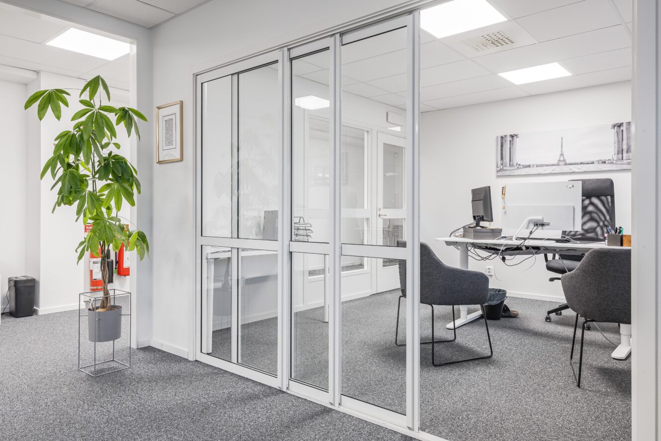 Ett modernt kontor med glasväggar, två skrivbord med datorer, två stolar och en stor krukväxt i förgrunden.