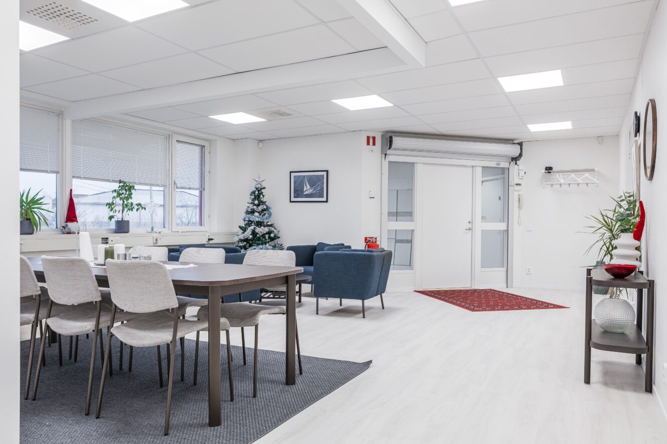 En modern, ljus kontorslounge med matbord, stolar, sittgrupp, krukväxter, en liten julgran och minimalistisk inredning.