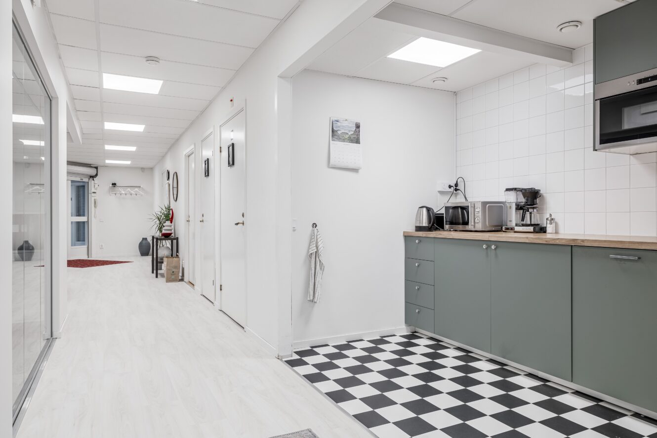 Ett modernt kontorskök med gröna skåp, kaffemaskiner och rutigt golv, bredvid en korridor med vita väggar och dörrar.