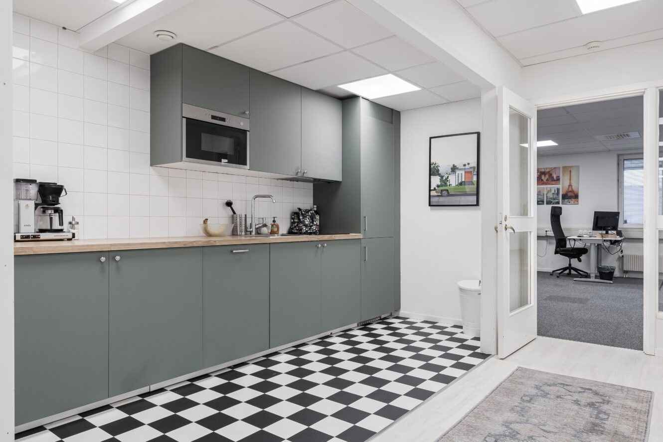 Modernt kontorskök med salviagröna skåp, svartvitrutigt golv, vitkaklat stänkskydd och öppen dörr som leder till ett kontorsområde med skrivbord och datorer.