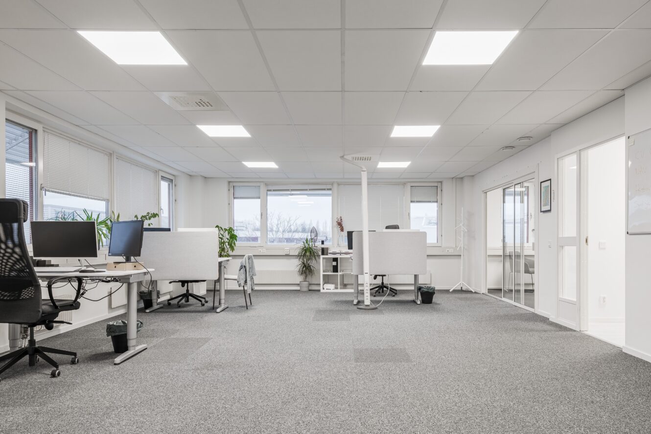 Ett modernt kontor med flera skrivbord, datorer, kontorsstolar och växter. Stora fönster ger naturligt ljus och utrymmet är organiserat med ett neutralt färgschema.