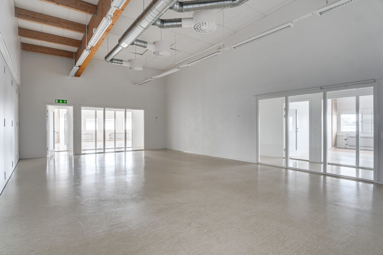 Rymligt, tomt rum med ljust golv, vita väggar, synliga rör i taket och stora glasdörrar som leder till angränsande rum.