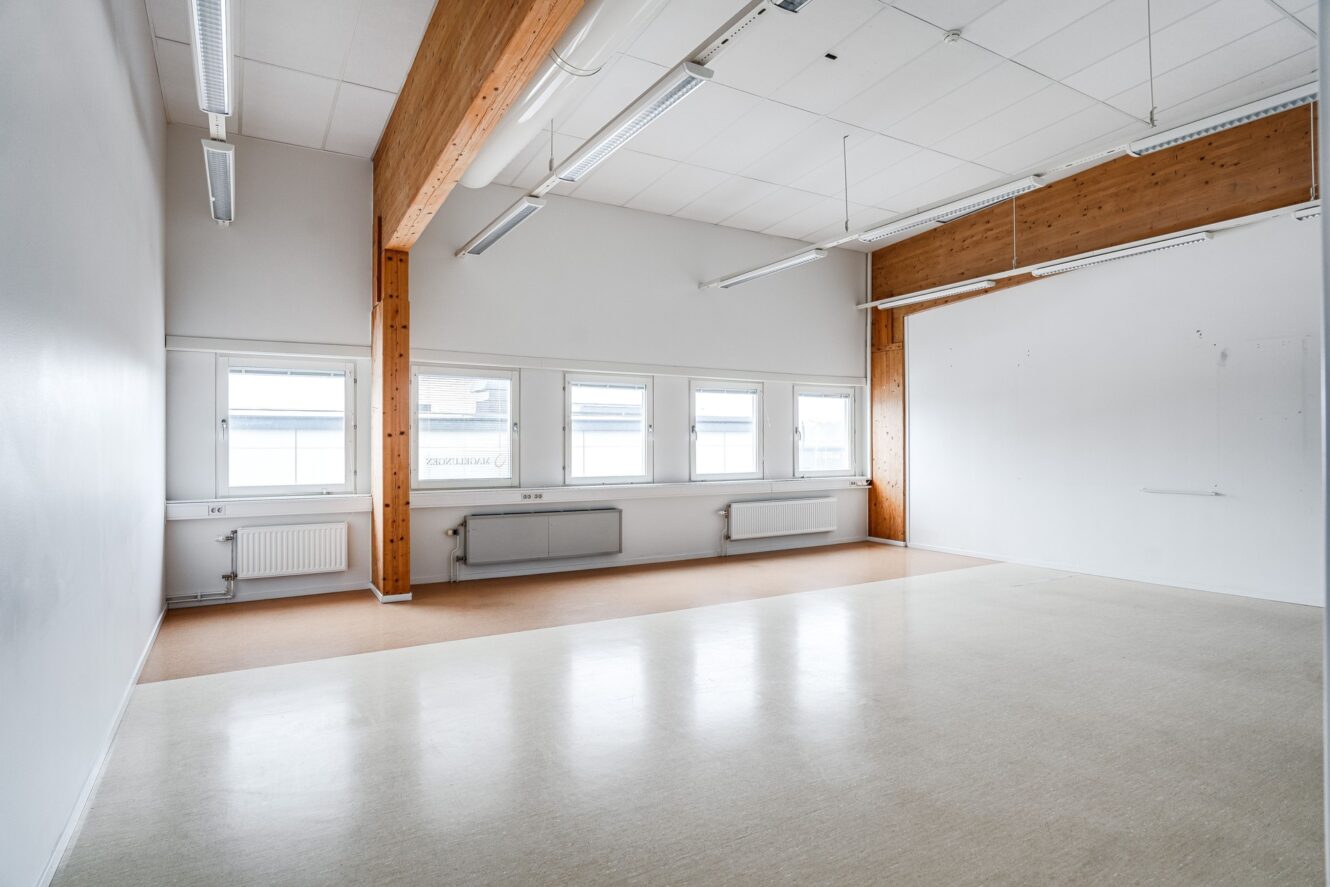 Tomt, väl upplyst rum med vita väggar, stora fönster, radiatorer, synliga träbjälkar, taklampor och ett beige linoleumgolv.