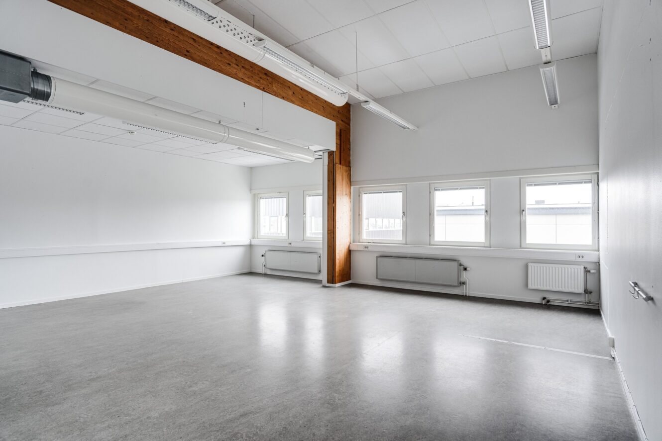 Tomt, omöblerat rum med vita väggar, stora fönster, betonggolv och synliga takbjälkar; lysrör och radiatorer syns.