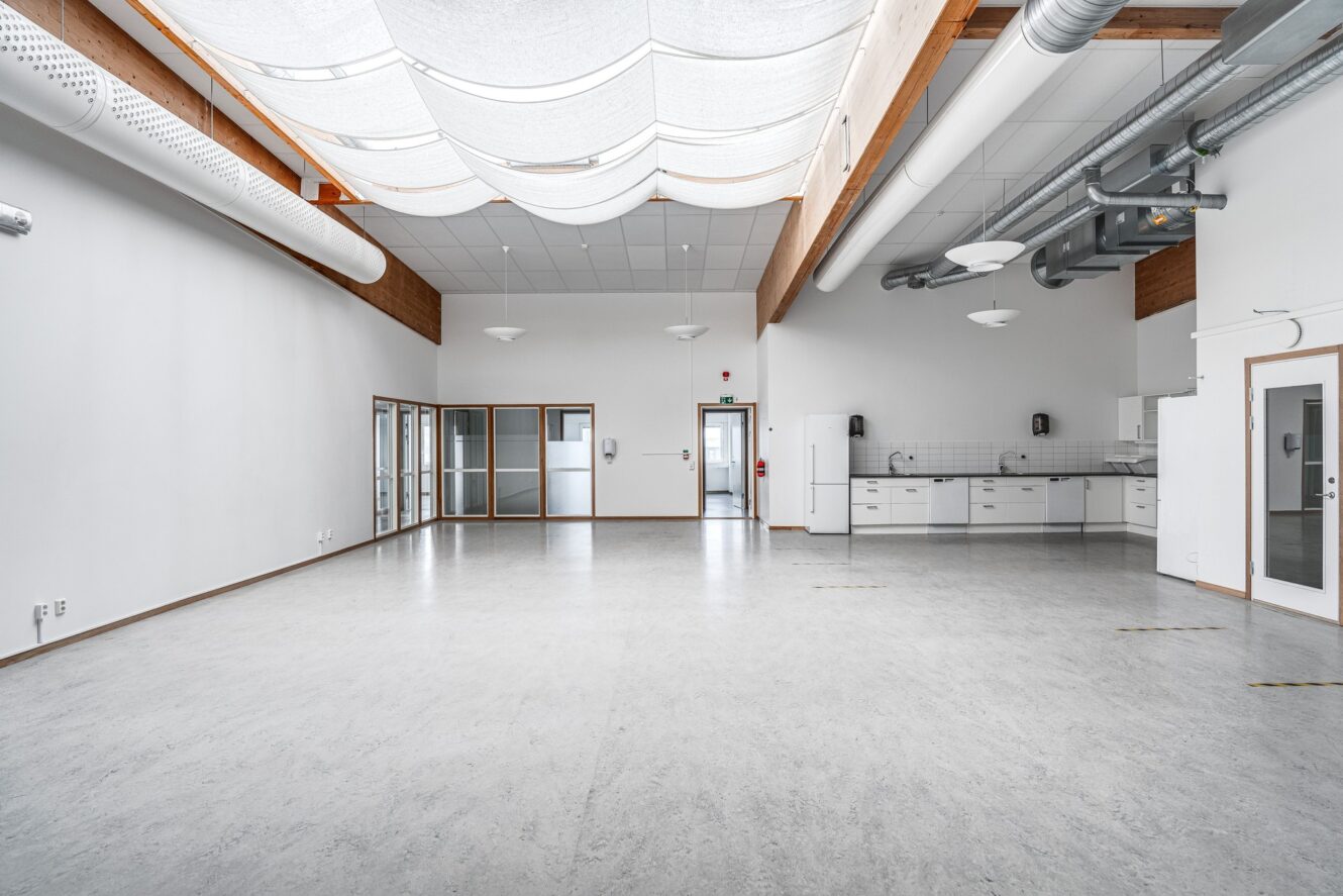 Rymligt, tomt rum med vita väggar, stora fönster, rör i taket, ett pentry på ena sidan och modern belysning.