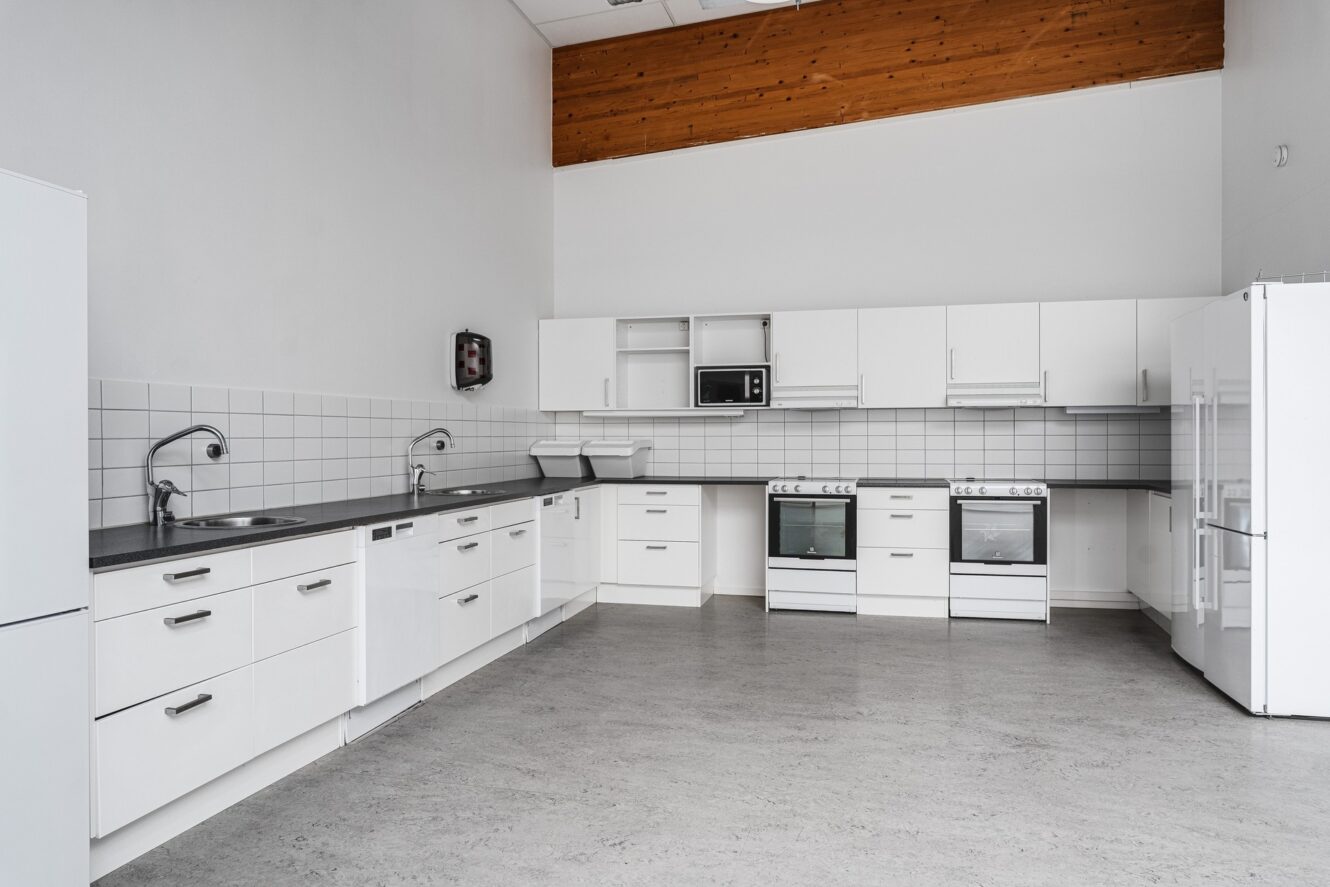 Ett modernt kök med vita skåp, två ugnar, två diskhoar, en mikrovågsugn och ett kylskåp, med grå bänkskivor och kaklat stänkskydd.