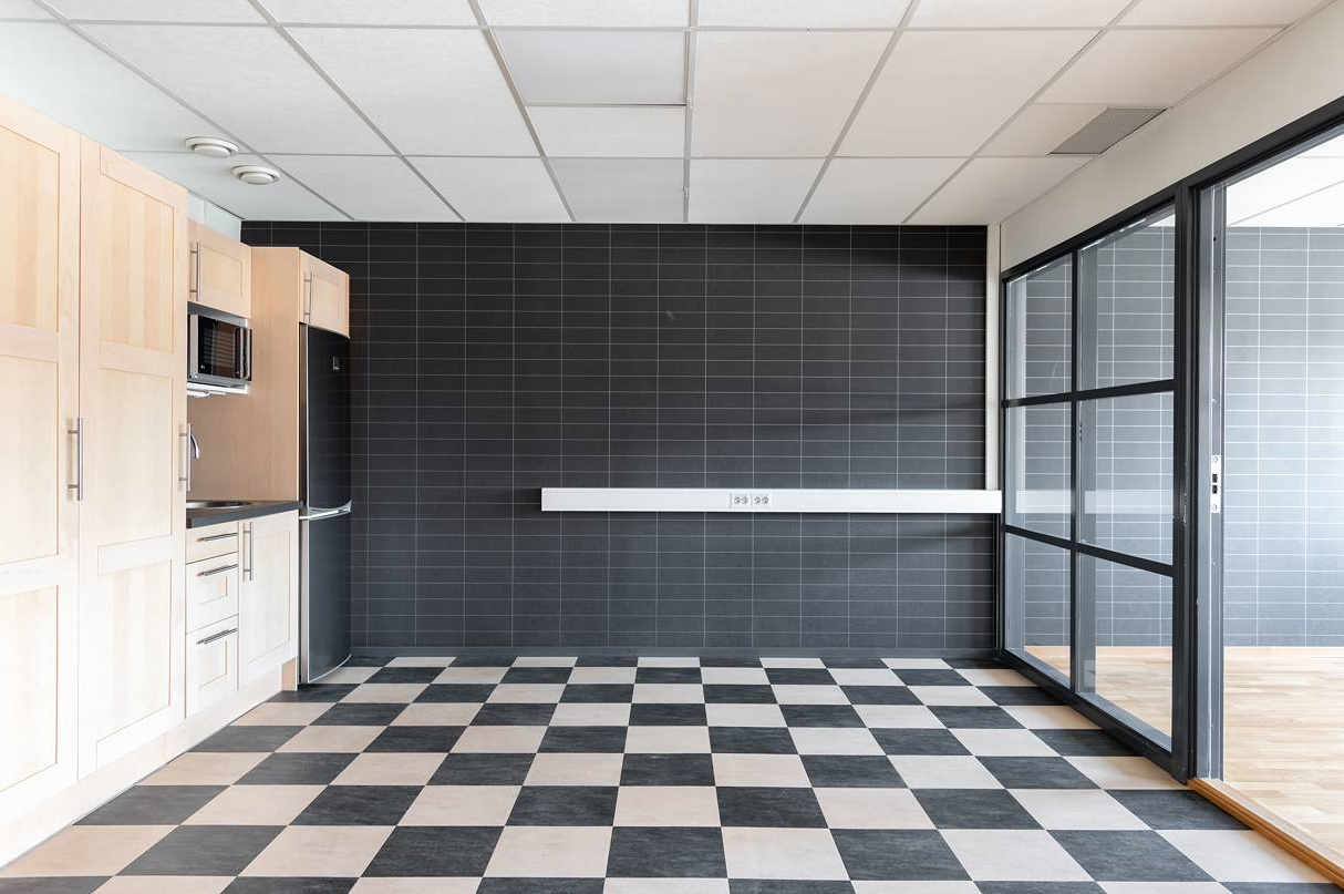 Modernt kök med skåp i ljust trä, vitvaror i rostfritt stål, svart kaklad vägg och ett svartvitrutigt golv intill en glasvägg.