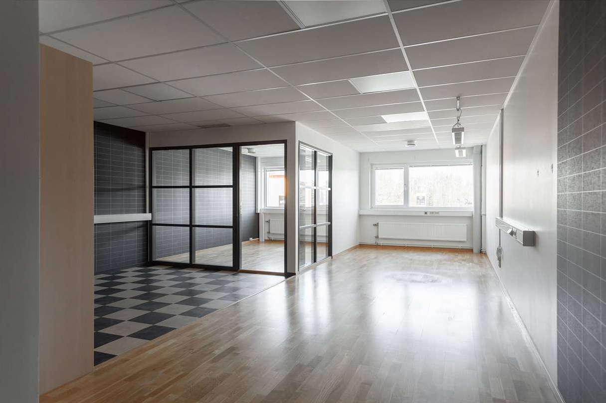 Ett modernt, tomt rum med stora fönster, ljust trägolv och en avgränsad del med glasväggar med svart ram och svartvitrutigt klinkergolv.