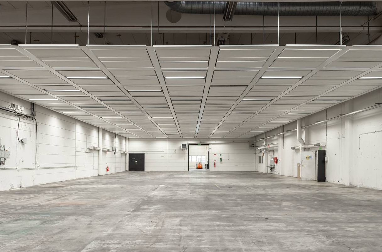 Stor, tom industrihall med vita väggar, synliga rör, mönstrat tak, betonggolv och stängda svarta dörrar längst bort.