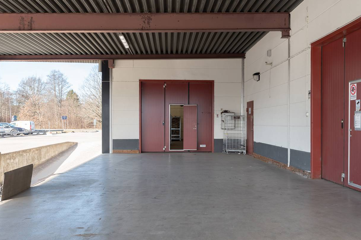 Överbyggd lastkaj med betonggolv, röda dubbeldörrar, trådvagn och utvändig vy över en parkeringsplats och träd i bakgrunden.