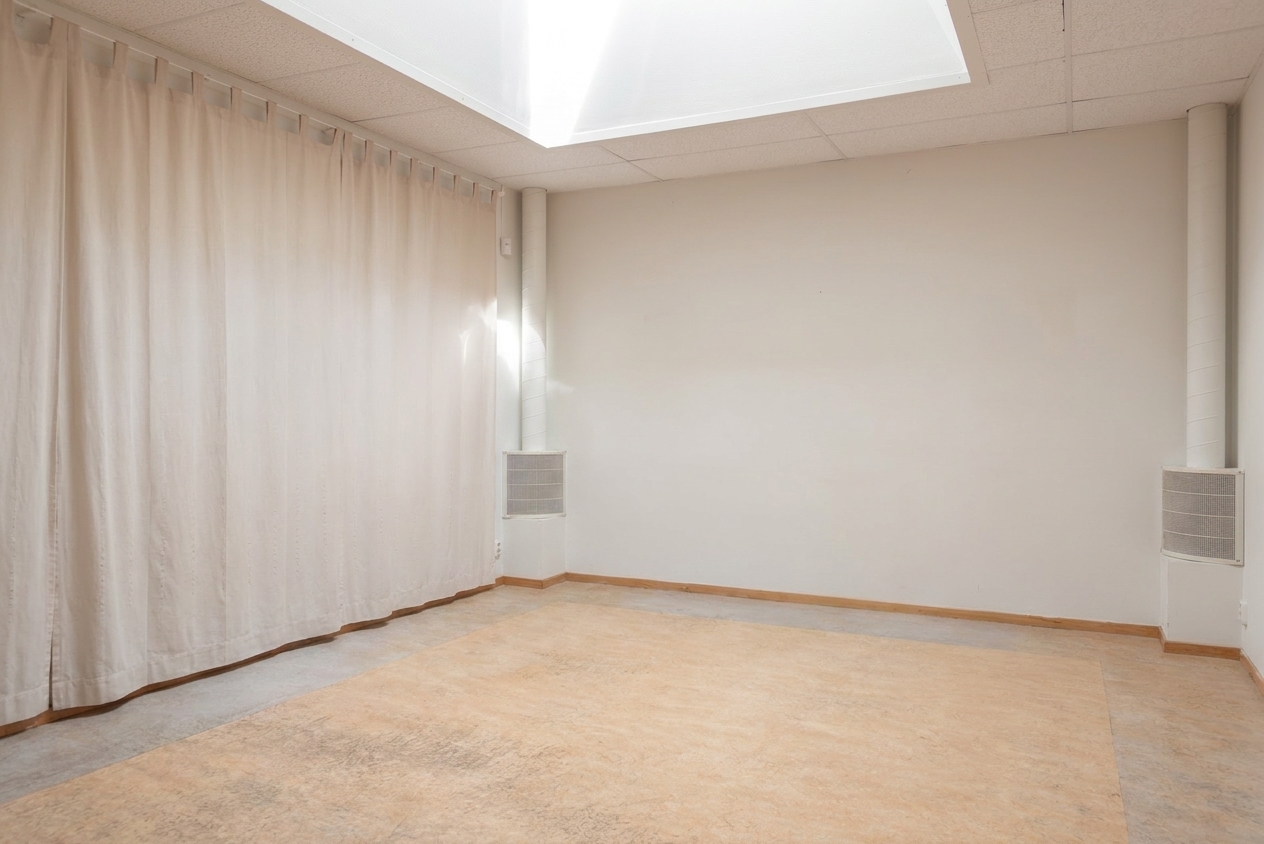Ett tomt beige rum med en ljus gardin på ena sidan, ett takfönster ovanför och enfärgade väggar med ventilationsöppningar i hörnen. Golvet är naket och rummet är väl upplyst.