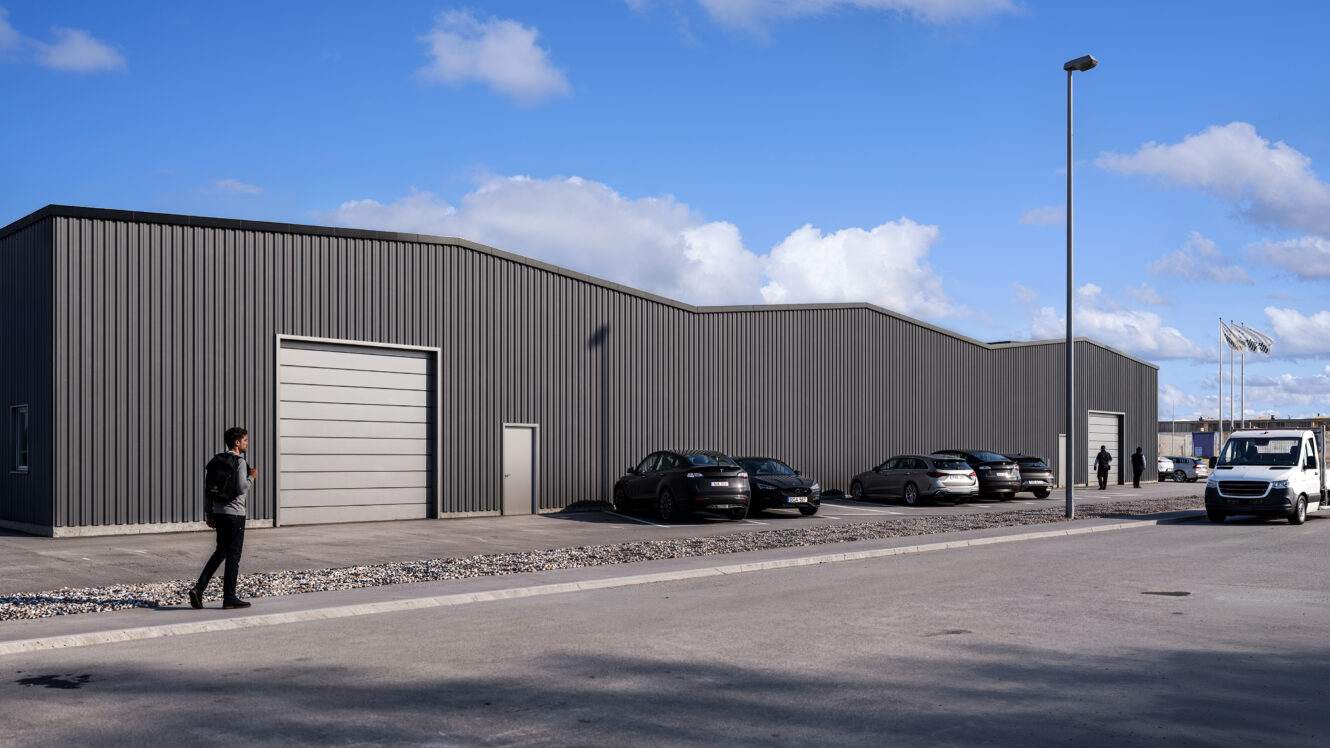 Industriella lagerbyggnader med grå metallfasader, flera parkerade bilar, en vit skåpbil och människor som går utomhus under en delvis molnig himmel.