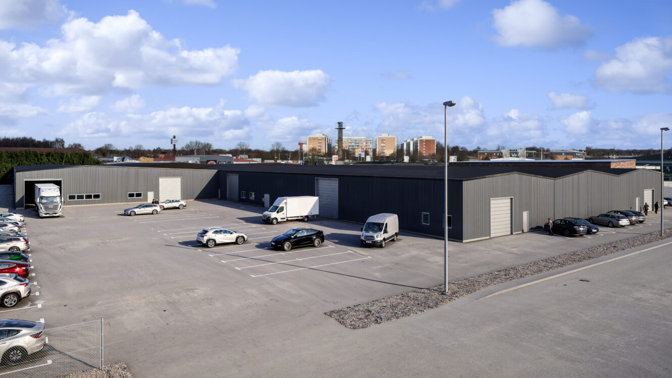 Ett stort, grått industriellt lagerkomplex med parkeringsplatser, flera bilar och leveransfordon under en delvis molnig himmel.