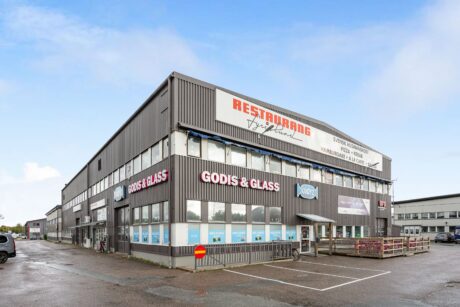 En grå industribyggnad med skyltar för en restaurang och en godis- och glassbutik, belägen på en parkeringsplats under en molnig himmel.