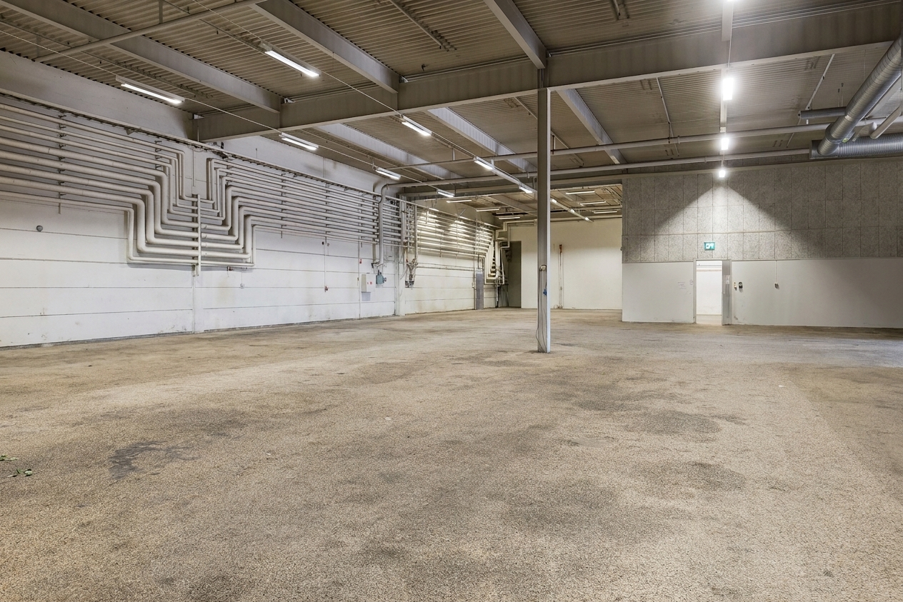 En stor tom lagerlokal med synliga rör på vita väggar, ett beige betonggolv, stödbalkar och lysrör i taket. En enda vit dörr syns på den bortre väggen.