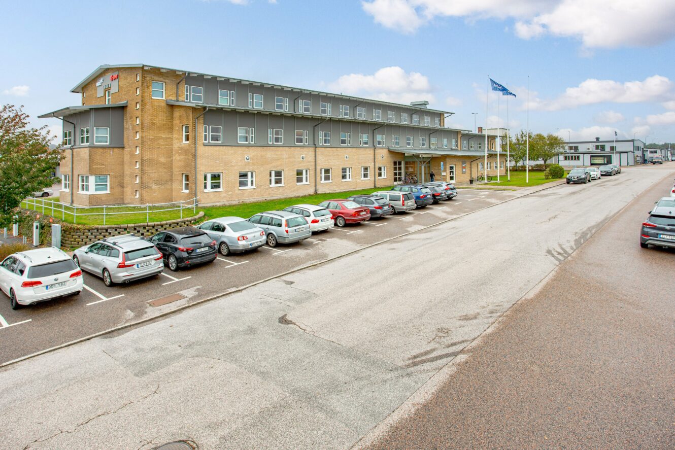 En trevåningsbyggnad i tegel och grått med parkerade bilar framför, flaggstänger och en molnig himmel över sig längs en asfalterad gata.