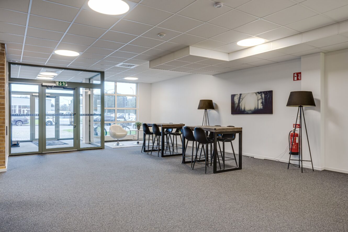 Modern kontorslobby med stora fönster, höga bord och stolar, två svarta golvlampor, en brandsläckare och ett inramat väggkonstverk; utomhusvy synlig genom glasdörrar.