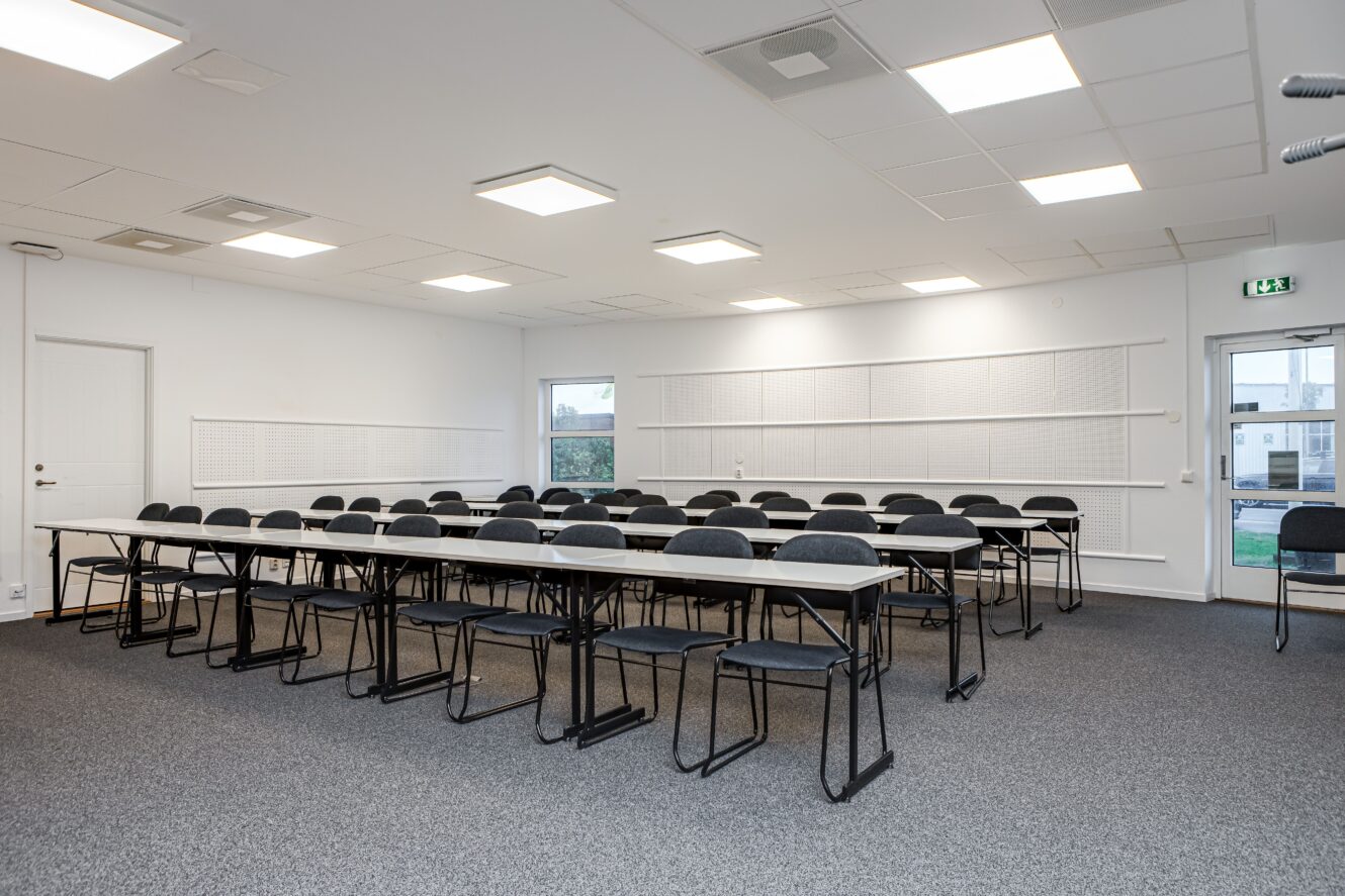 Ett modernt, tomt klassrum med rader av svarta stolar och vita bord placerade framför stora fönster och en whiteboardtavla.
