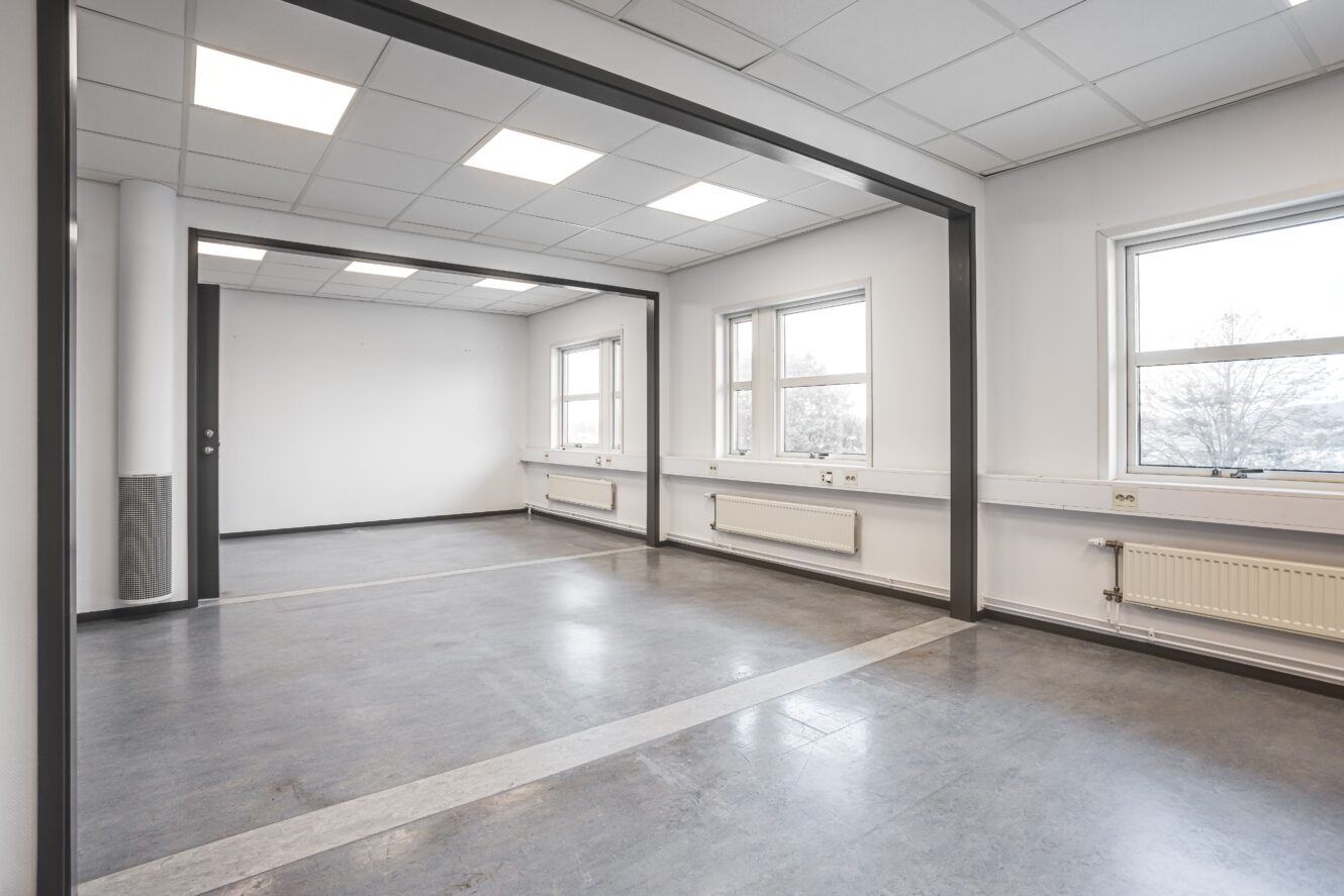 Tomt kontorsutrymme med stora fönster, vita väggar och grått golv. Rummet har lysrörsbelysning och synliga radiatorer under fönstren.