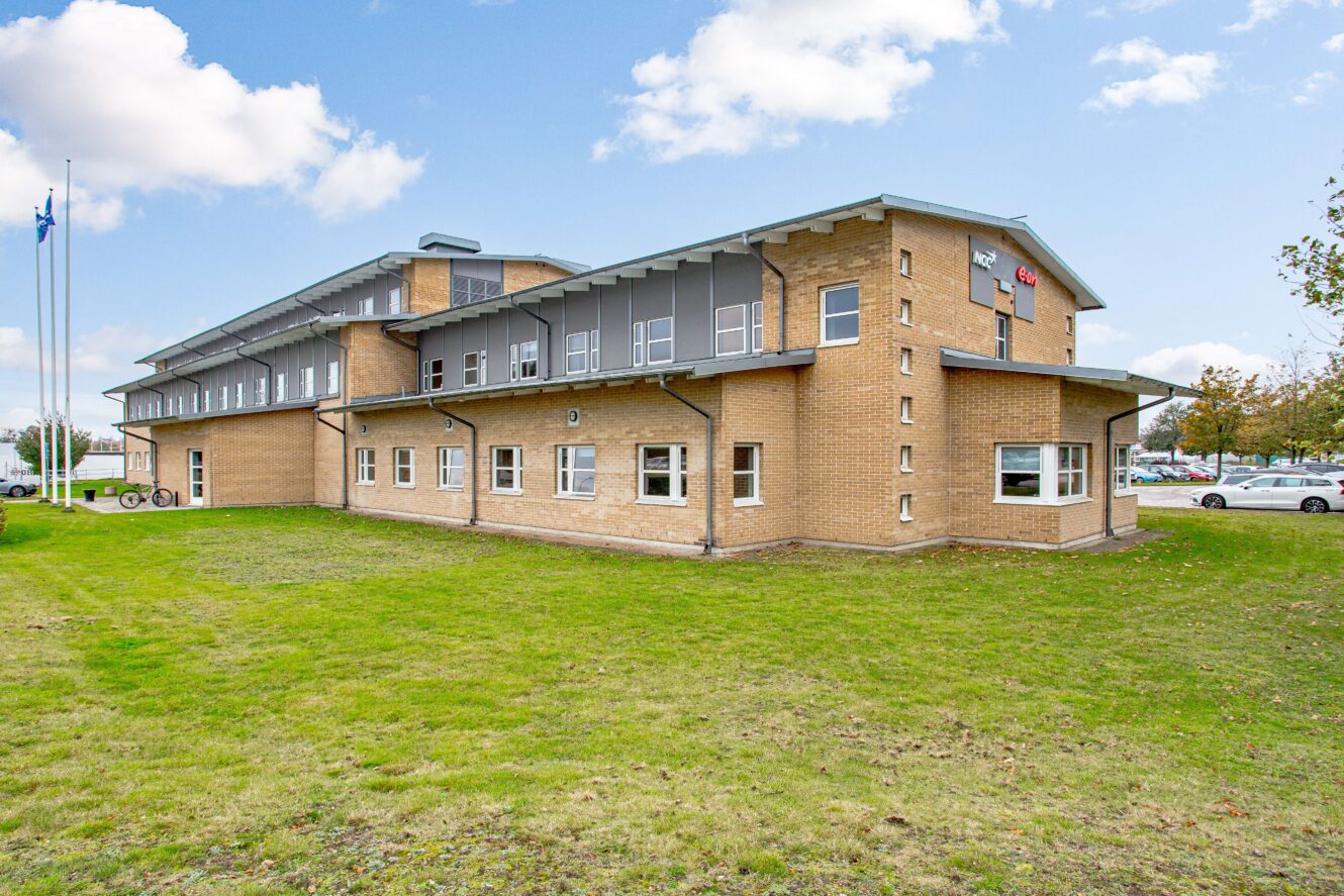 En tvåvånings tegelbyggnad med flera fönster, ett grått tak och en gräsmatta framför, under en delvis molnig himmel.