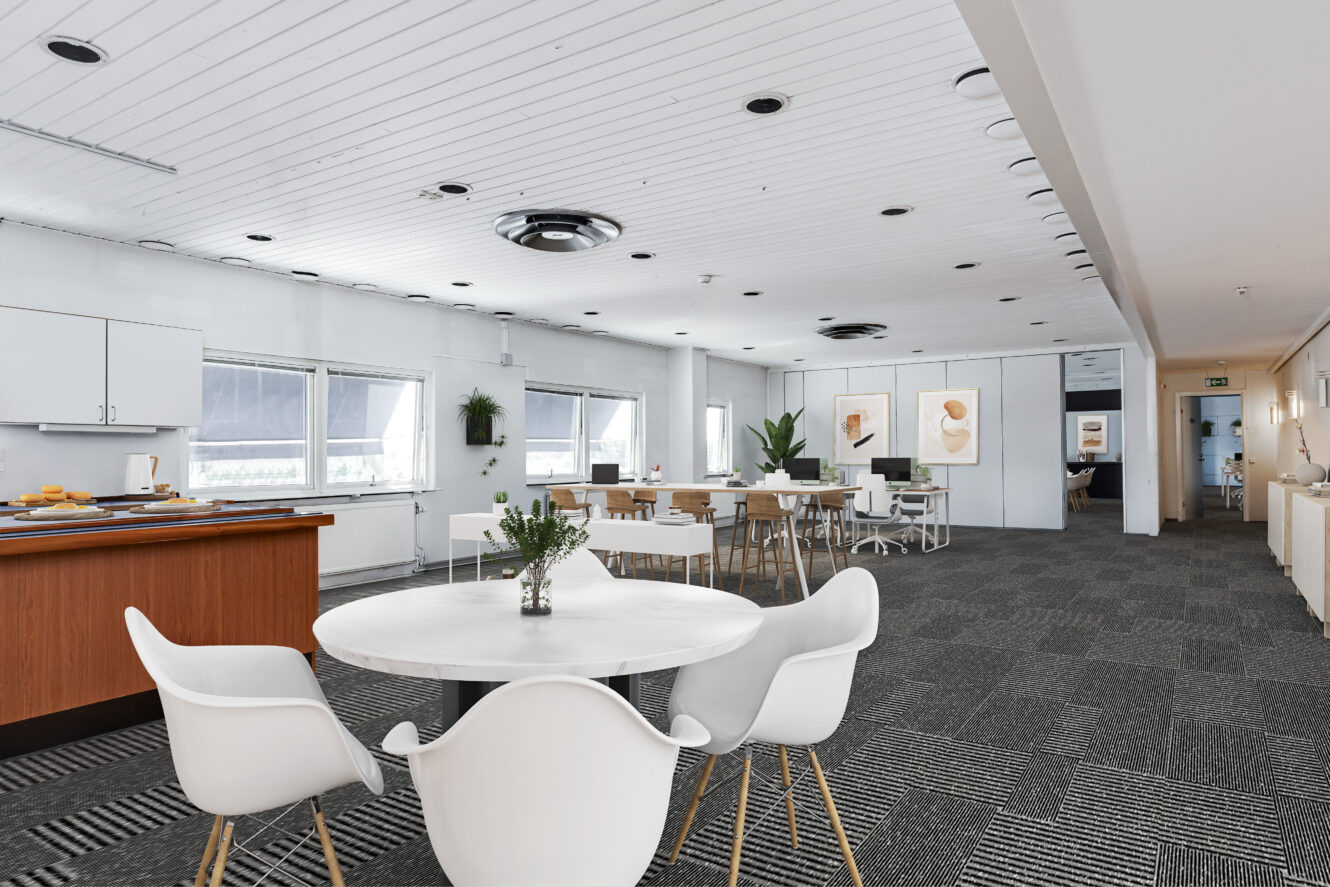 Moderna kontorslokaler med runda bord och vita stolar, stora fönster, grå heltäckningsmatta och väggkonst, med ett pentry till vänster.