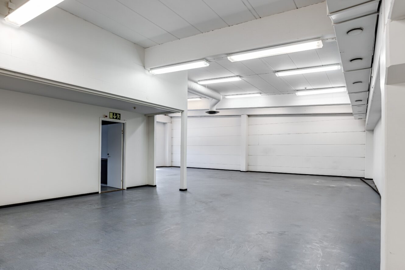 Tomt, väl upplyst industrirum med vita väggar, grått golv, lysrör i taket, en stödpelare, ventilationsrör och en öppen dörr med en utgångsskylt.