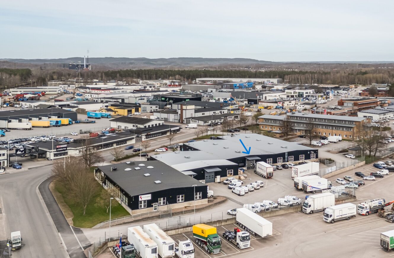 Flygbild över ett industriområde med flera lagerlokaler, parkerade lastbilar och bilar. En blå pil pekar på en specifik byggnad nära mitten av bilden.