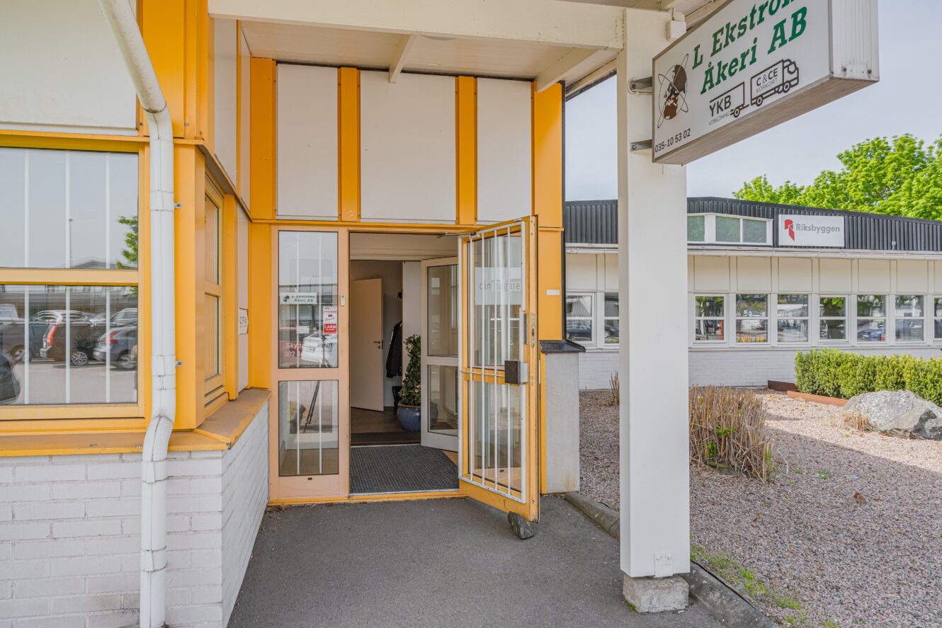 Entré till en kommersiell byggnad med gula och vita väggar, en öppen metallgrind och en skylt med texten L Ekström Åkeri AB. I bakgrunden syns buskar och parkerade bilar.