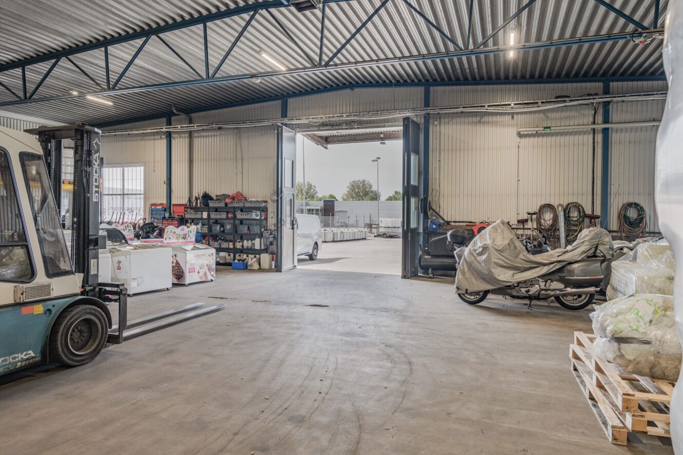 Lagerinredning med en gaffeltruck till vänster, en täckt motorcykel till höger, hyllor med varor och stora öppna dörrar som leder ut till en parkeringsplats.