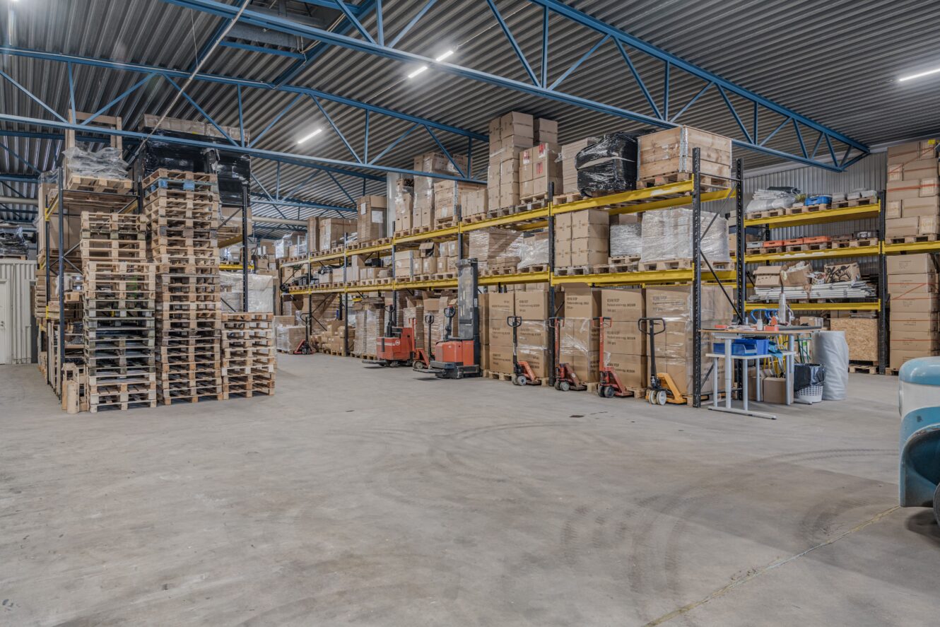 Lagerlokal med hyllor fyllda med lådor och pallar, pallbockar och utrustning som syns i en organiserad, industriell miljö.