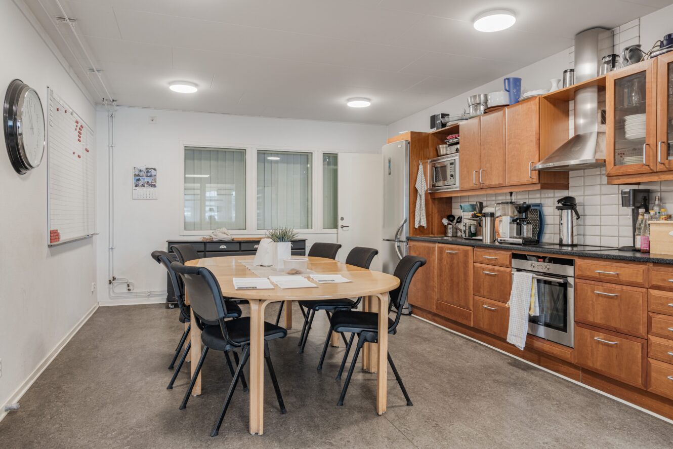 Ett fikarum med ett träbord och sex svarta stolar, ett pentry med vitvaror och skåp samt en whiteboardtavla på väggen.