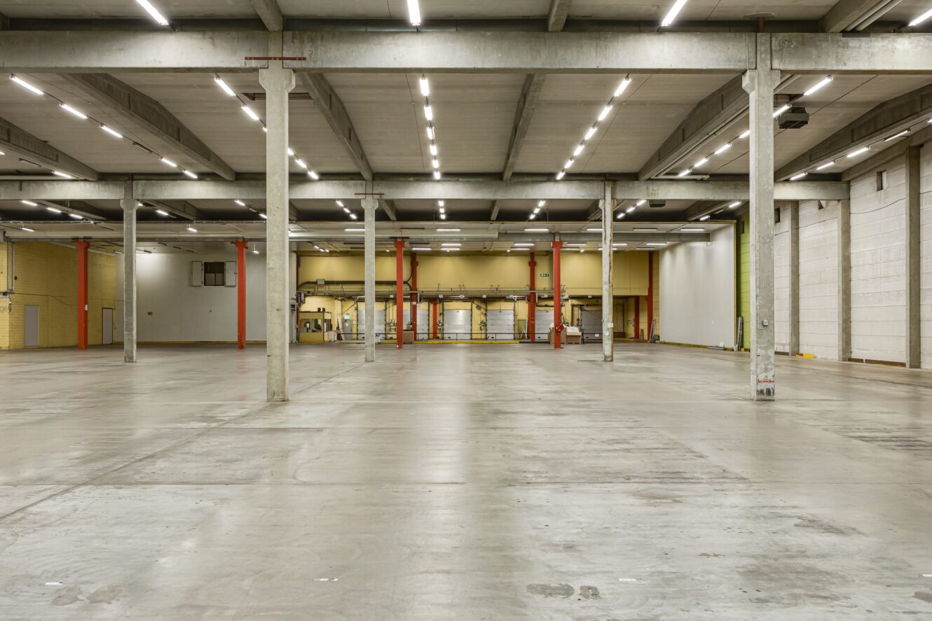 Stor tom lagerlokal med betonggolv, högt i tak, lysrörsbelysning och bärande pelare. Lastkajer och dörrar syns i bortre änden av utrymmet.