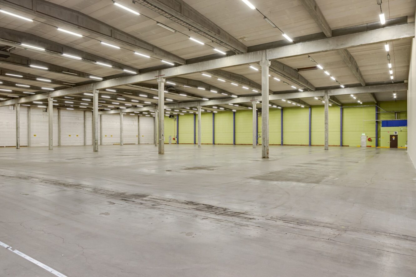 Stor tom lagerlokal med betonggolv, högt i tak, synliga balkar, lysrörsbelysning och ljusgrön vägg i bakgrunden.