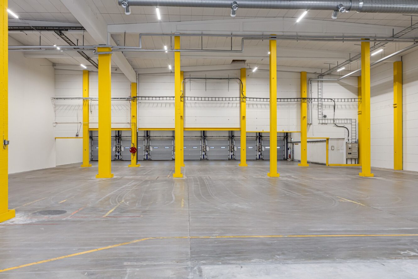 Interiör av en tom lagerlokal med gula stödpelare, takbelysning och flera lastkajsdörrar längs bakväggen.