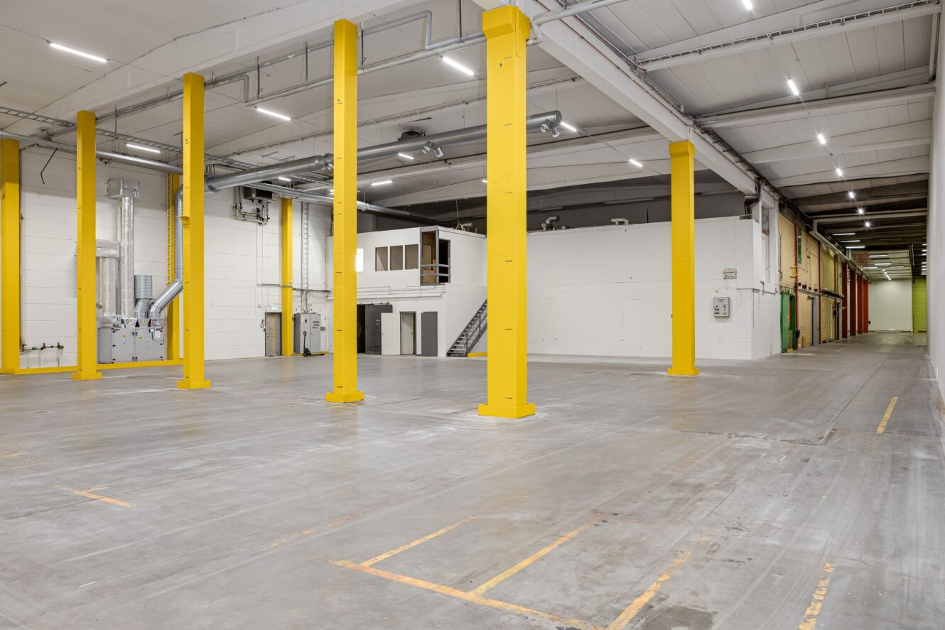 Stor tom lagerlokal med gula stödpelare, betonggolv, vita väggar och takbelysning.