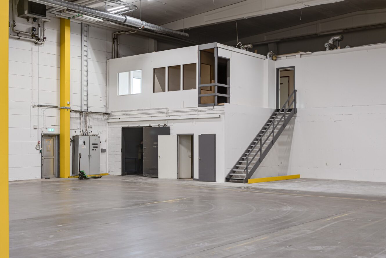 Tomt industriellt lagerutrymme med betonggolv, vita väggar, flera dörrar, en trappa som leder till ett högt beläget kontorsrum och synliga rör i taket.