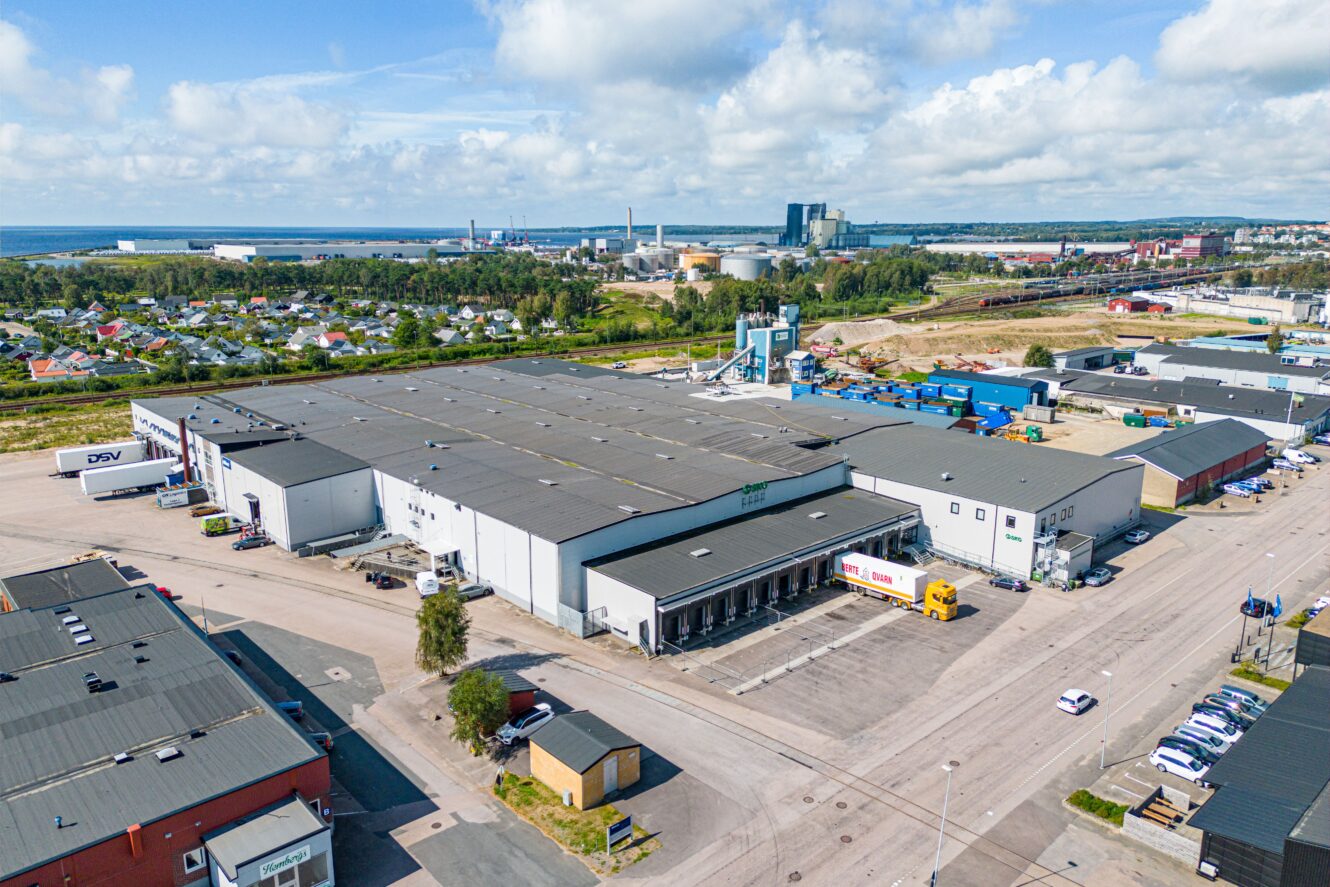 Flygfoto över ett industriellt lagerkomplex med lastkajer, lastbilar och omgivande byggnader; bostadsområde och industrianläggningar i bakgrunden.