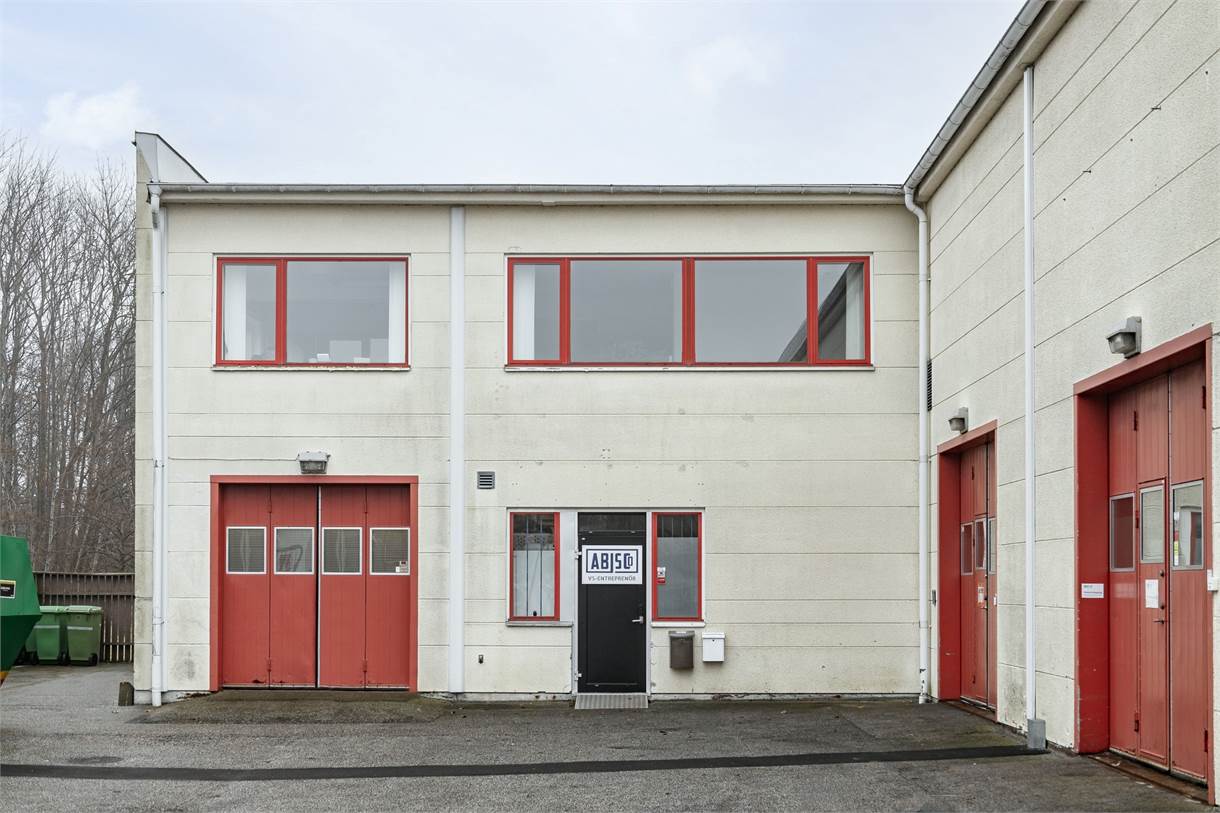 En industribyggnad i två våningar med beige väggar och rödkarmade fönster och dörrar; en skylt med texten ABISEJO finns på entrédörren.