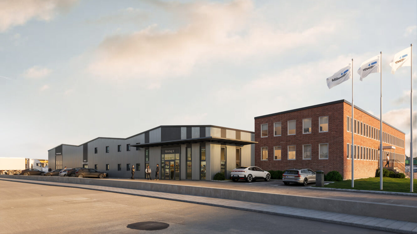 En modern industribyggnad med en blandning av tegel- och metallfasader, flera parkerade bilar och tre vita företagsflaggor en klar dag.