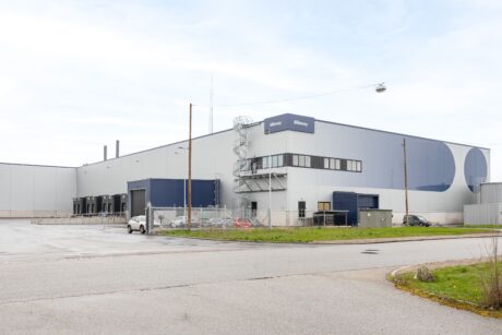 En stor grå och blå industriell lagerbyggnad med en parkeringsplats, kedjelänkstaket och några fordon utanför en molnig dag.