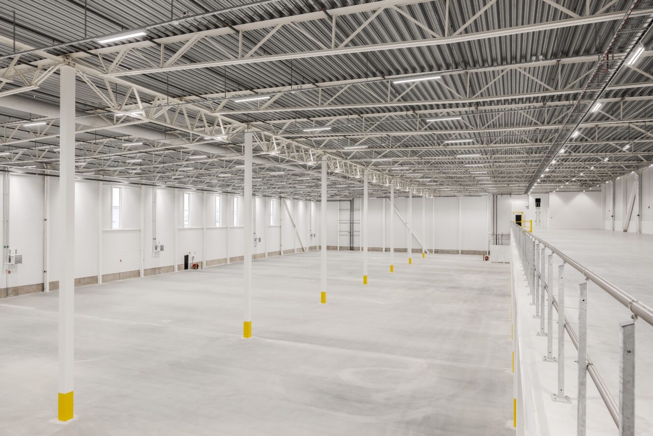 Stor, tom industrilokal med högt i tak, vita väggar, synliga bjälkar och gulrandiga pelare; ett entresolplan löper längs höger sida.