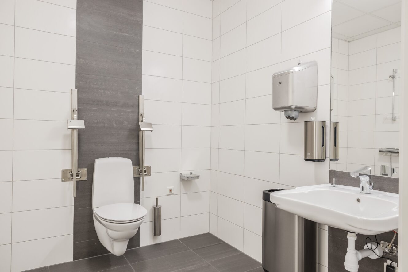 Tillgänglig toalett med vägghängd toalett, stödskenor, vitkaklade väggar, handfat med spegel, pappershandduksdispenser och papperskorg.