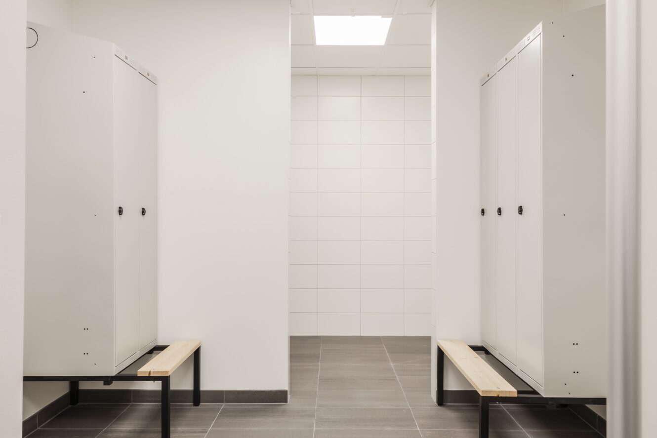 Ett rent, modernt omklädningsrum med vita skåp, klinkergolv, bänkar i ljust trä och vitkaklade väggar under en taklampa.