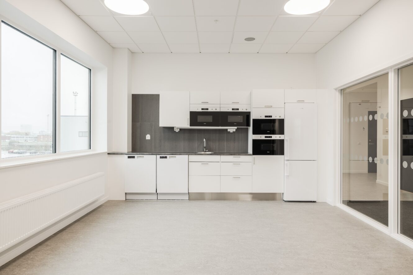 Ett modernt, minimalistiskt kök med vita skåp, vitvaror i rostfritt stål, stort fönster och glasvägg.