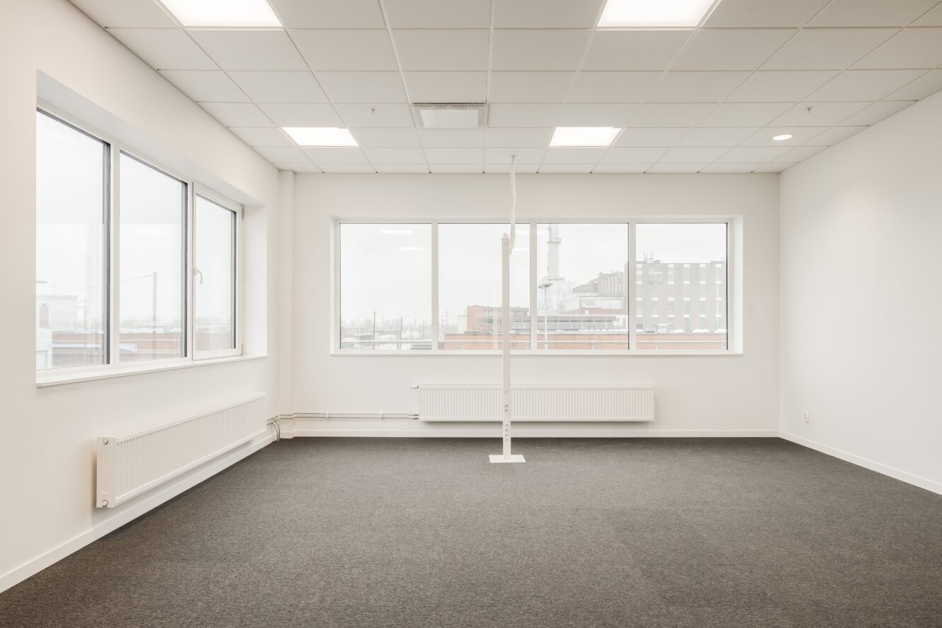 Tomt kontorsutrymme med stora fönster, vita väggar, kaklat tak med lysrör och grå heltäckningsmatta. Byggnader syns genom fönstren.