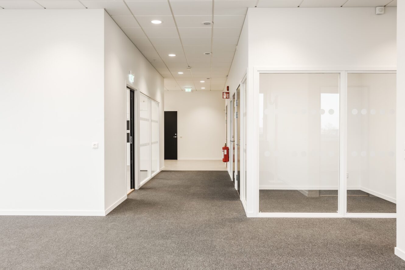 En modern, tom kontorskorridor med vita väggar, grå matta, kontor med glasväggar till höger och stängda dörrar längs korridoren.
